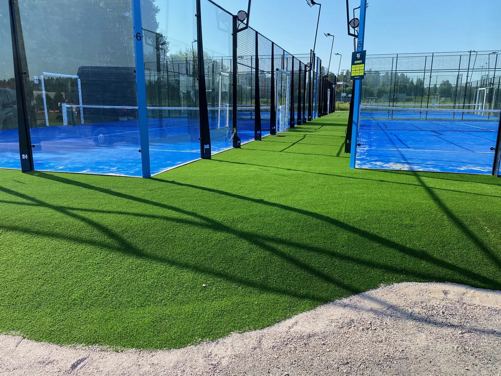 Tekonurmi padelkenttien välisillä käytävillä, taustalla sinisiä kenttiä lasiseinien takana.