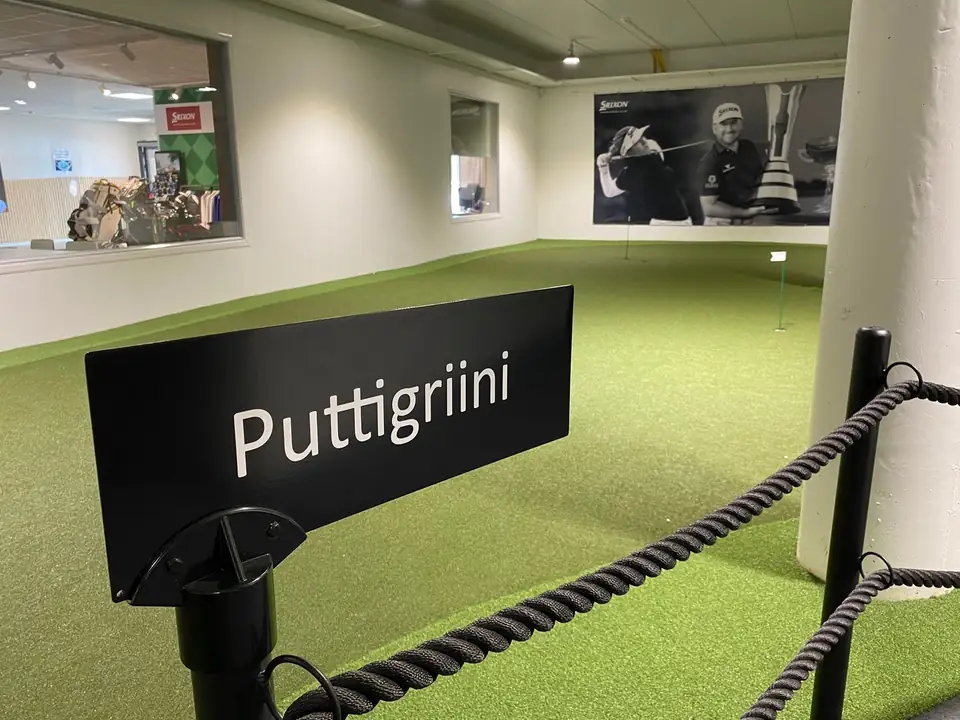 Puttigriini-kyltti alakerran viheriön köysiaidassa, taustalla golfaajien kuvia seinällä.