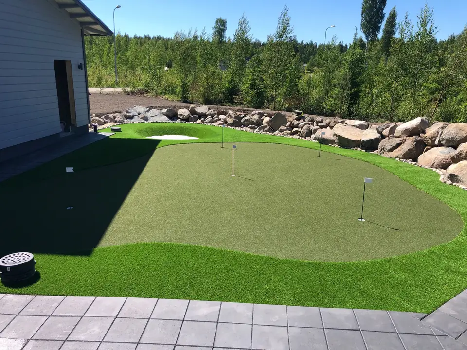 Kolmen lipun puttiviheriö talon kulmalla, takana bunkkeri ja luonnonkivistä tehty reunus.